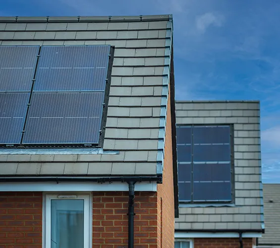 Close-up of two houses with grey tiled roofs and solar panels installed, set against a background of blue sky and light clouds.