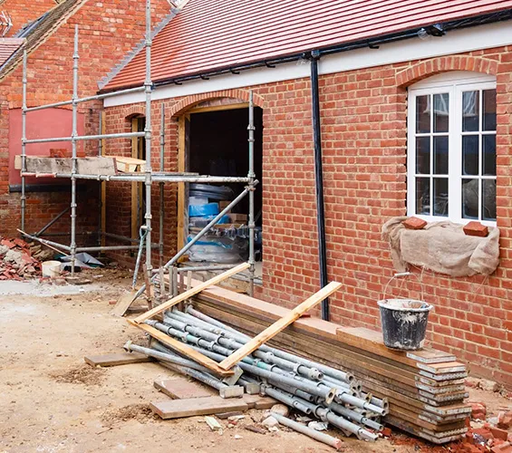 A brick building under construction with scaffolding, a pile of metal pipes, wooden planks, and a black bucket in front.