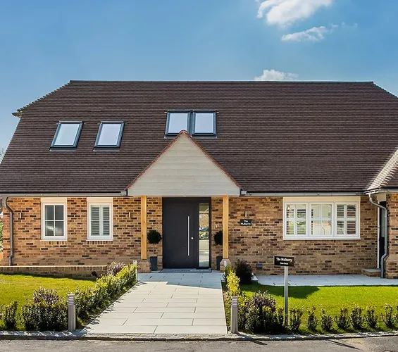 A brick house with a dark roof and large windows, a central path leading to the front door, and a short hedge outlining the lawn.