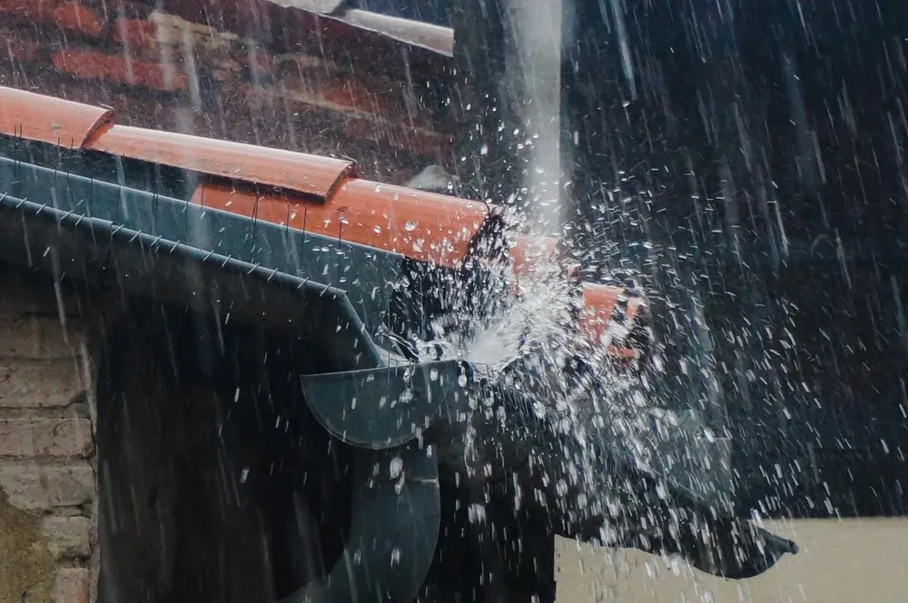 Rainwater overflowing and splashing from a clogged roof gutter during heavy rainfall.