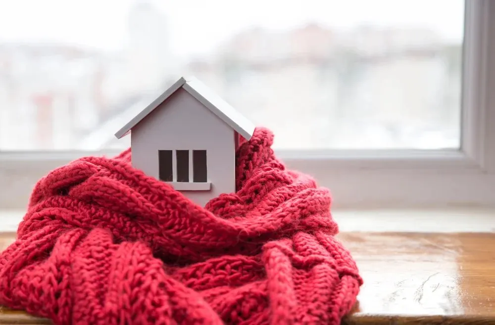 A small model house wrapped in a knitted red scarf sits on a windowsill with an out-of-focus cityscape in the background.