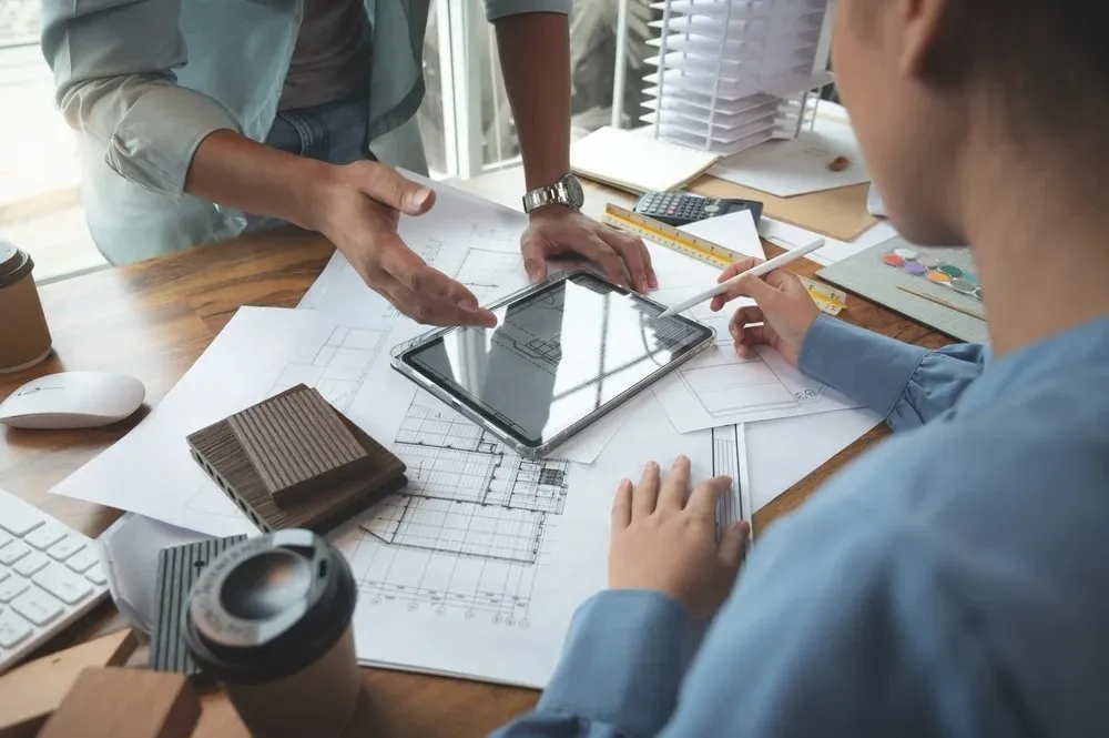 Two people in a meeting discuss architectural plans on a tablet, surrounded by blueprints, samples, and coffee cups on a desk.