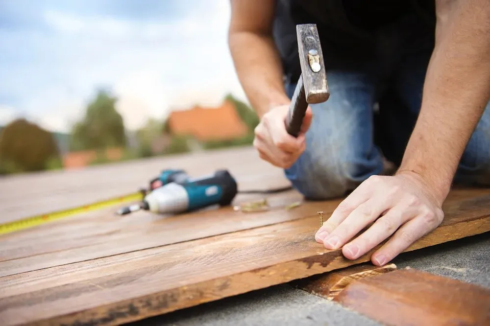 Person hammering a nail into a wooden plank, with tools including a measuring tape and drill resting nearby.