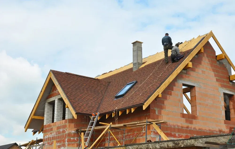 Two construction workers are installing roofing shingles on a partially built brick house under a cloudy sky.