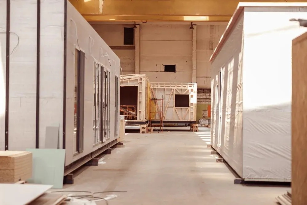 Inside a factory, prefabricated house sections are in various stages of assembly, lined up in a spacious production area.