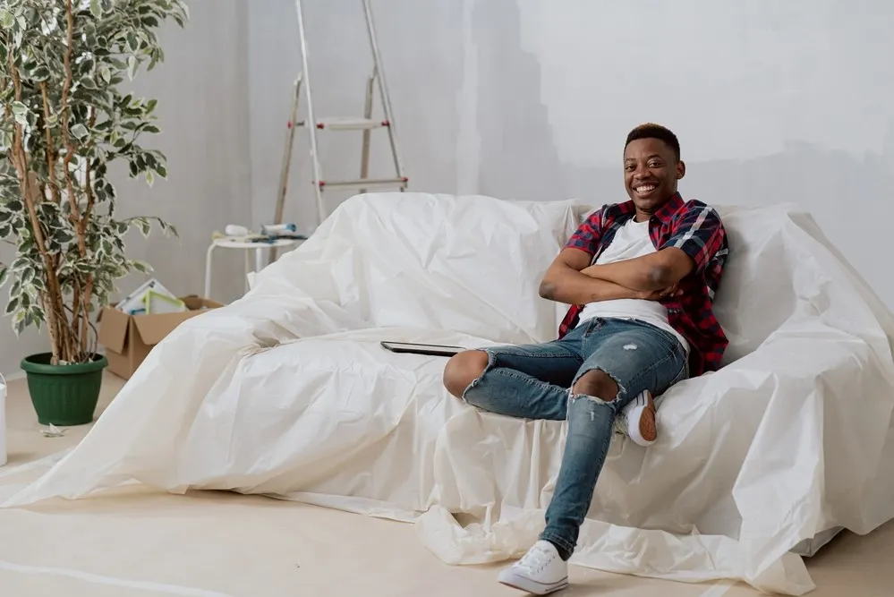 Person seated on a plastic-covered sofa, smiling. Ladder and plant in background, room appears to be in progress of renovation or painting.