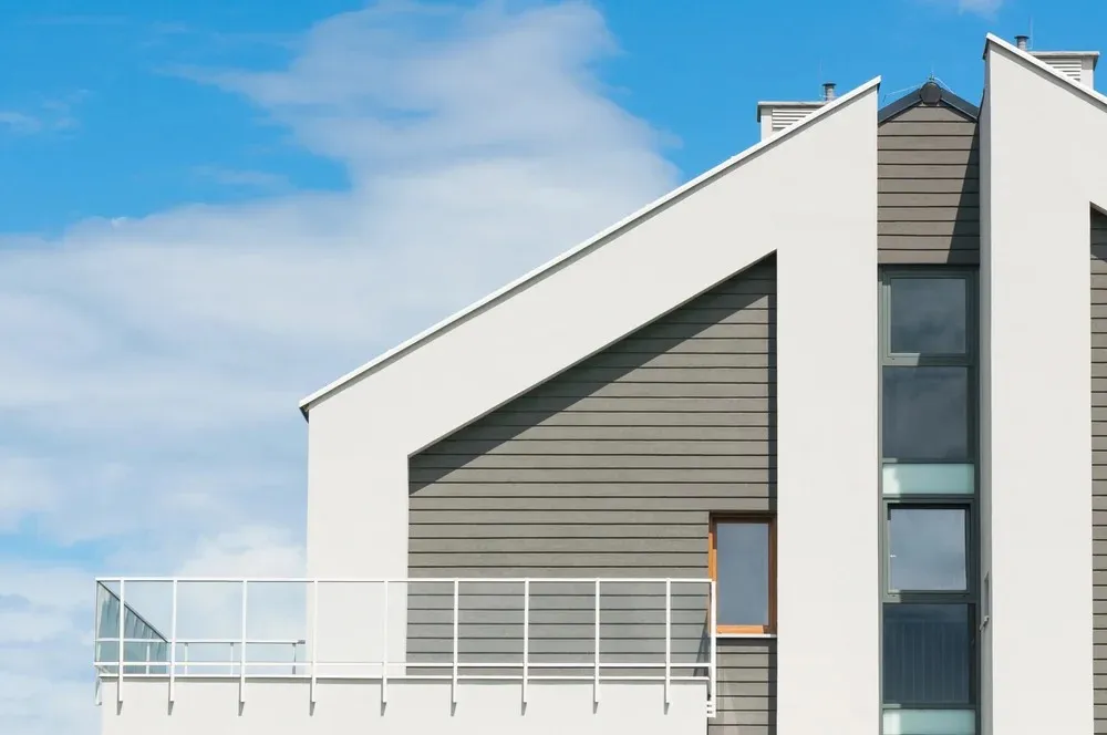 Modern building facade with geometric design, featuring gray horizontal siding, white trim, and a glass balcony under a blue sky.