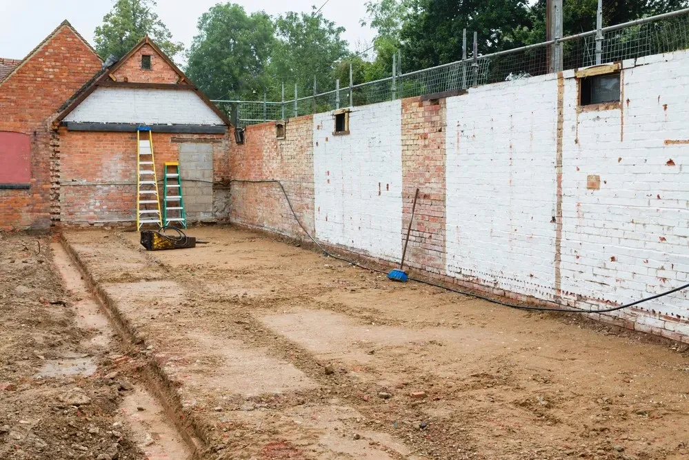 Empty construction site with exposed soil and foundation, surrounding brick walls, a ladder, and construction equipment present.