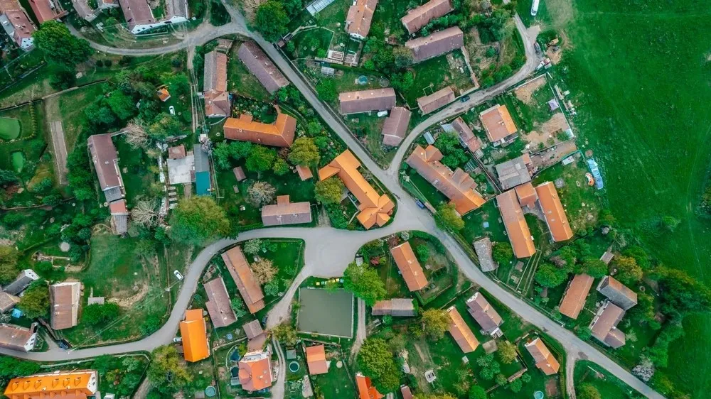 Aerial view of a rural village with clusters of houses with orange roofs, green fields, and winding roads.