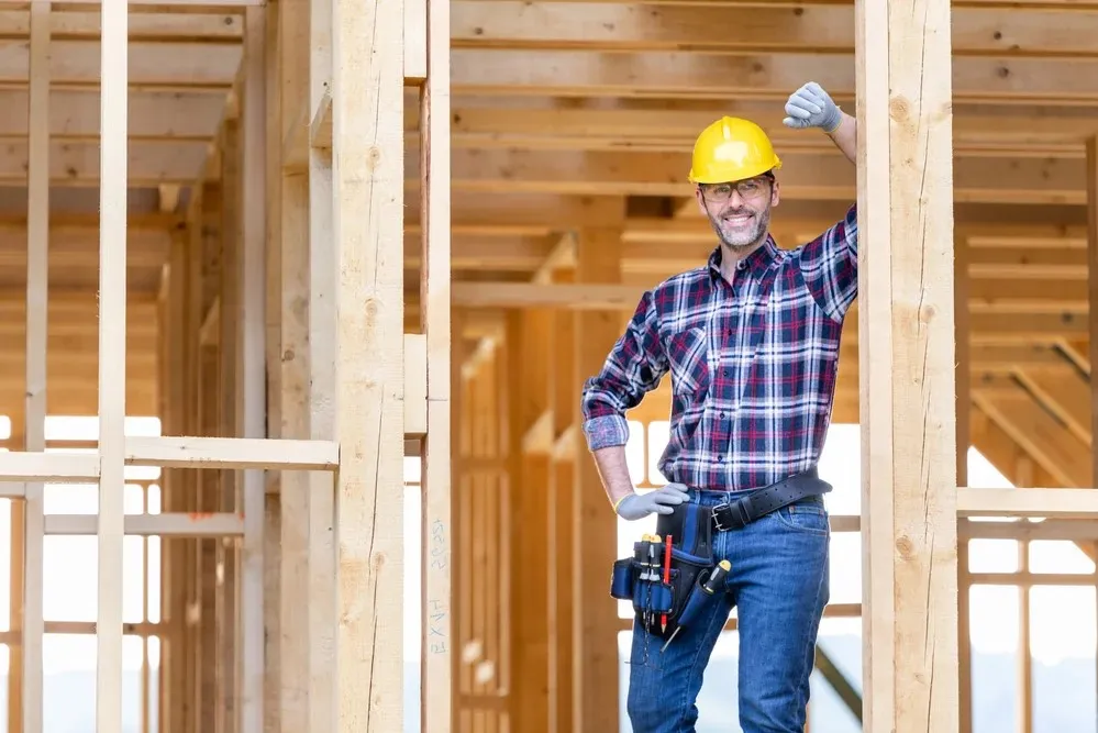 A construction worker in a safety helmet and plaid shirt stands inside a wooden building frame, smiling and raising one arm.