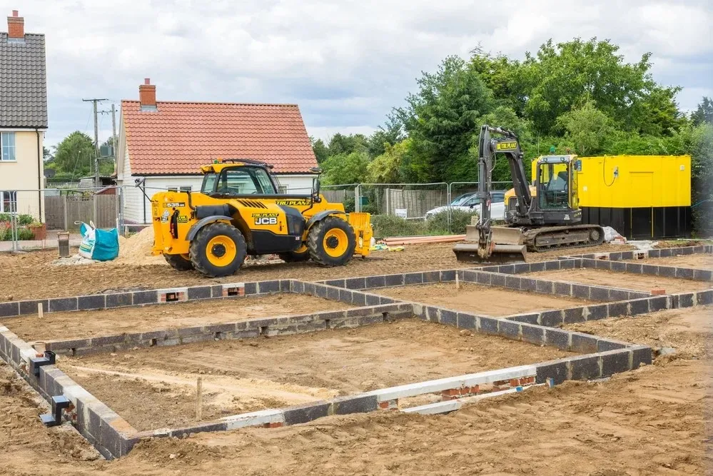 Construction site with a yellow JCB telehandler, an excavator, and foundation work in progress, surrounded by houses and trees.