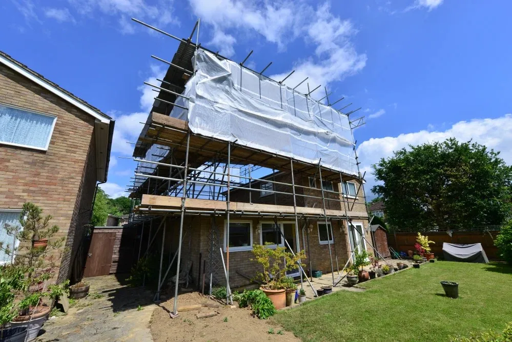 A house under renovation is covered in scaffolding and white sheeting, with a garden and plants in the foreground.