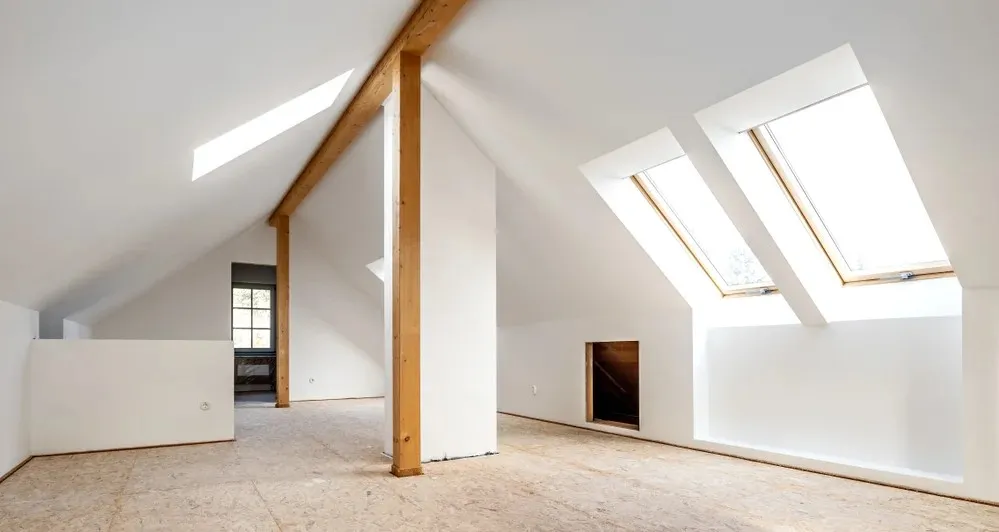 A spacious, unfinished attic with slanted ceilings, wooden beams, and skylights, featuring a large window at the far end.