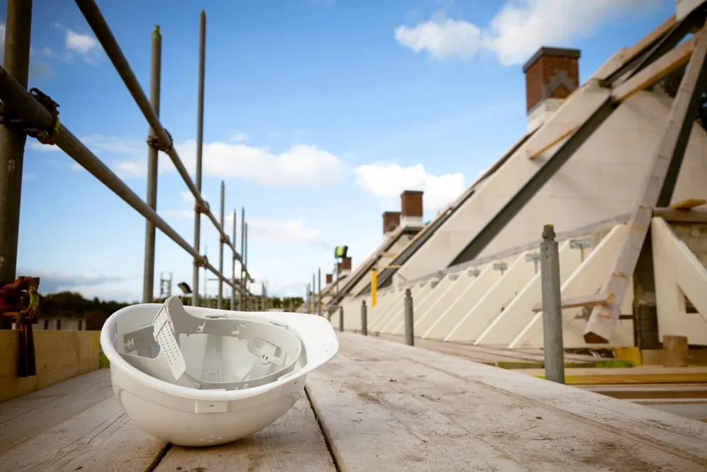 A white hard hat rests on wooden scaffolding at a construction site with building frameworks and a clear sky in the background.