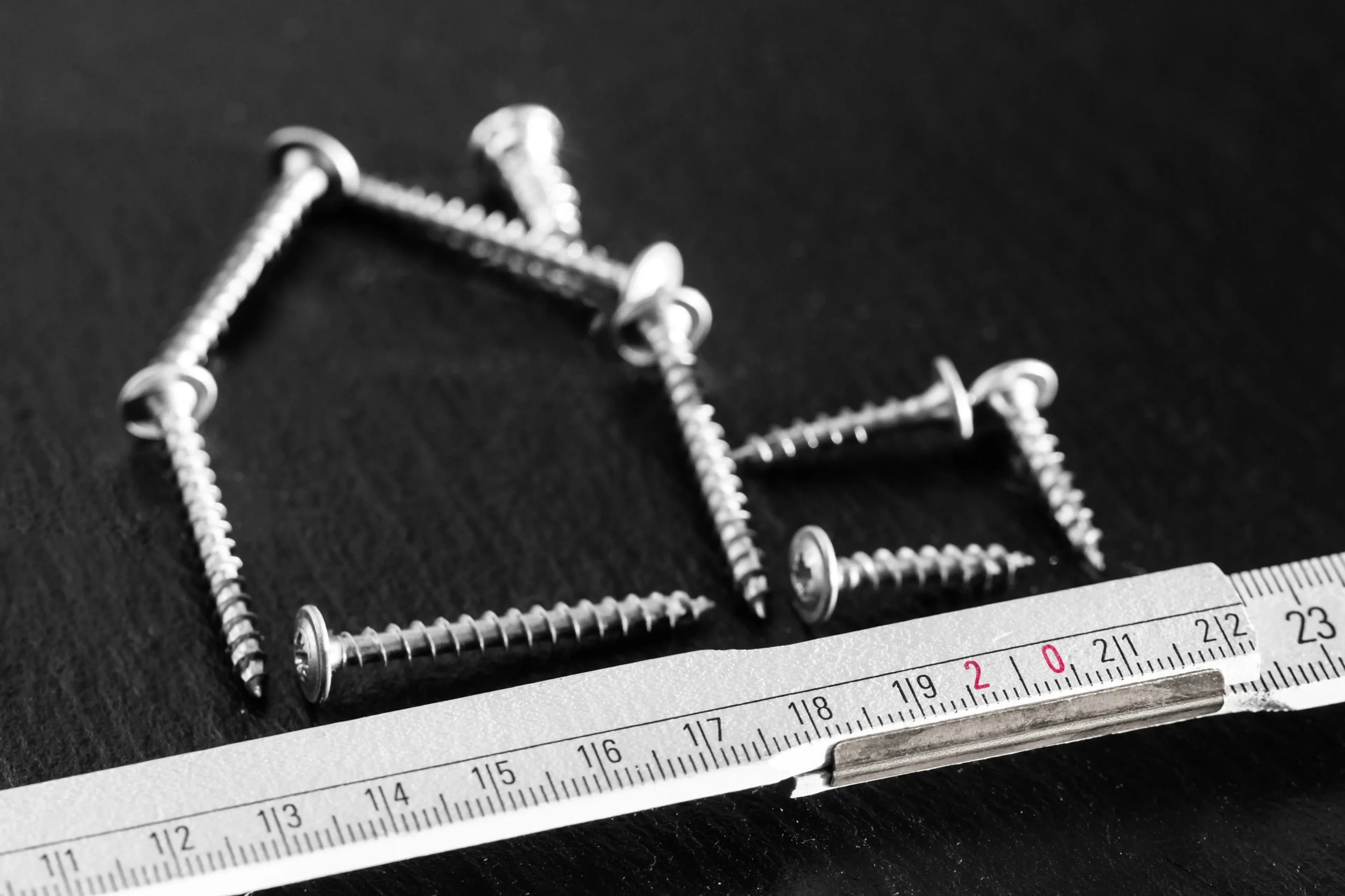 Screws arranged in the shape of a house next to a measuring tape on a black surface.