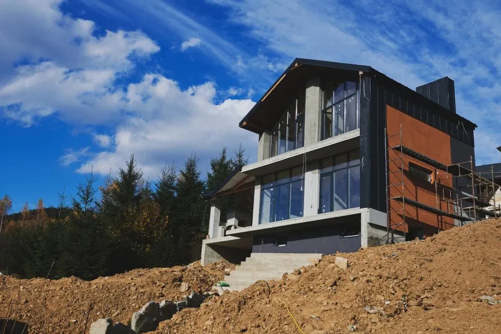 A modern two-story house under construction on a sloped terrain with scaffolding visible on one side, surrounded by dirt and trees.