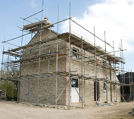 A two-story stone building under construction, surrounded by scaffolding on all sides, with a partly cloudy sky in the background.