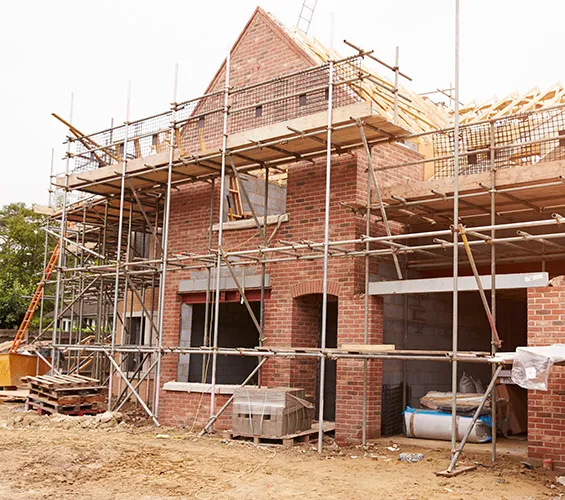 A two-story brick house under construction, with scaffolding around the structure and building materials on the ground.