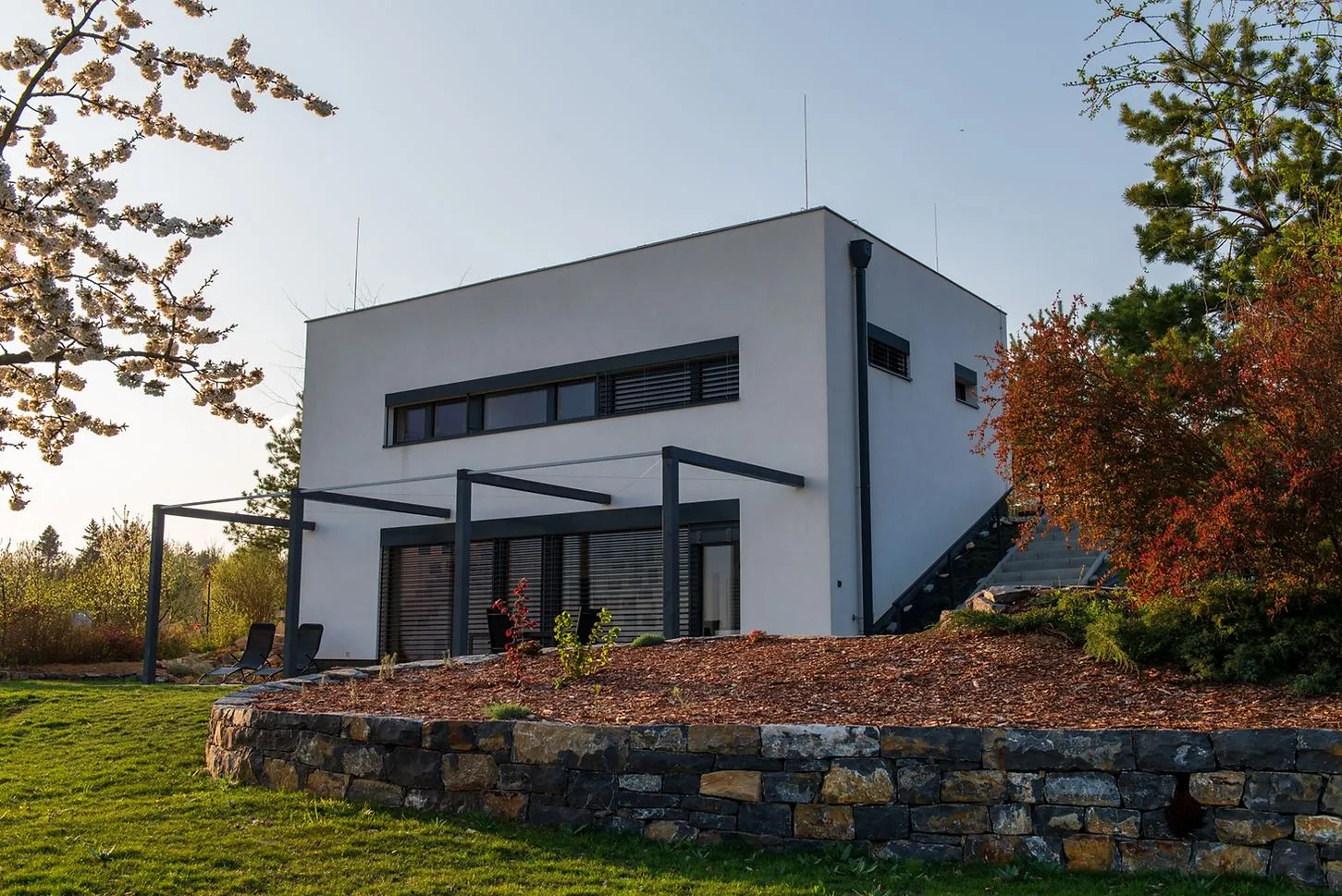 A modern, self-built two-story white house and a metal pergola designed to passive house standards, surrounded by greenery.