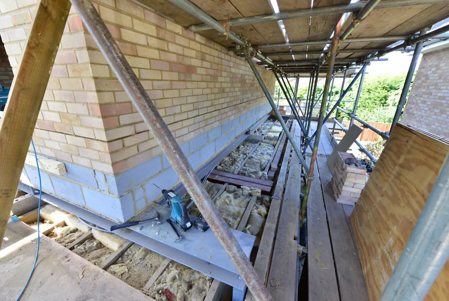 A construction site with scaffolding adjacent to a brick wall, featuring tools, building materials, and insulation on the ground.