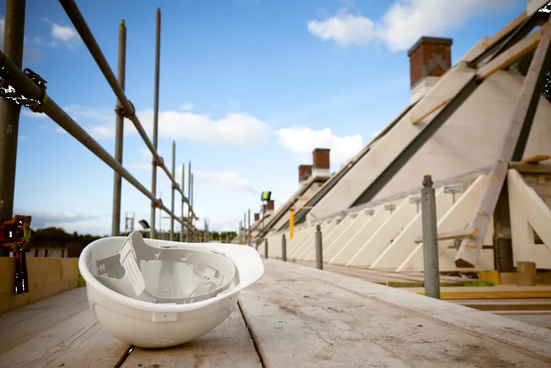 A white hard hat rests on a wooden scaffold at a construction site, amidst unfinished structures under a clear blue sky.