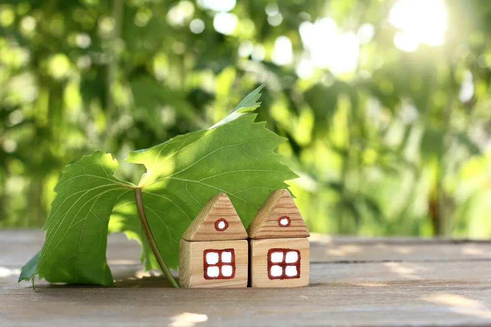 Two small wooden house models with red windows and a large green leaf, set against blurred greenery in the background.